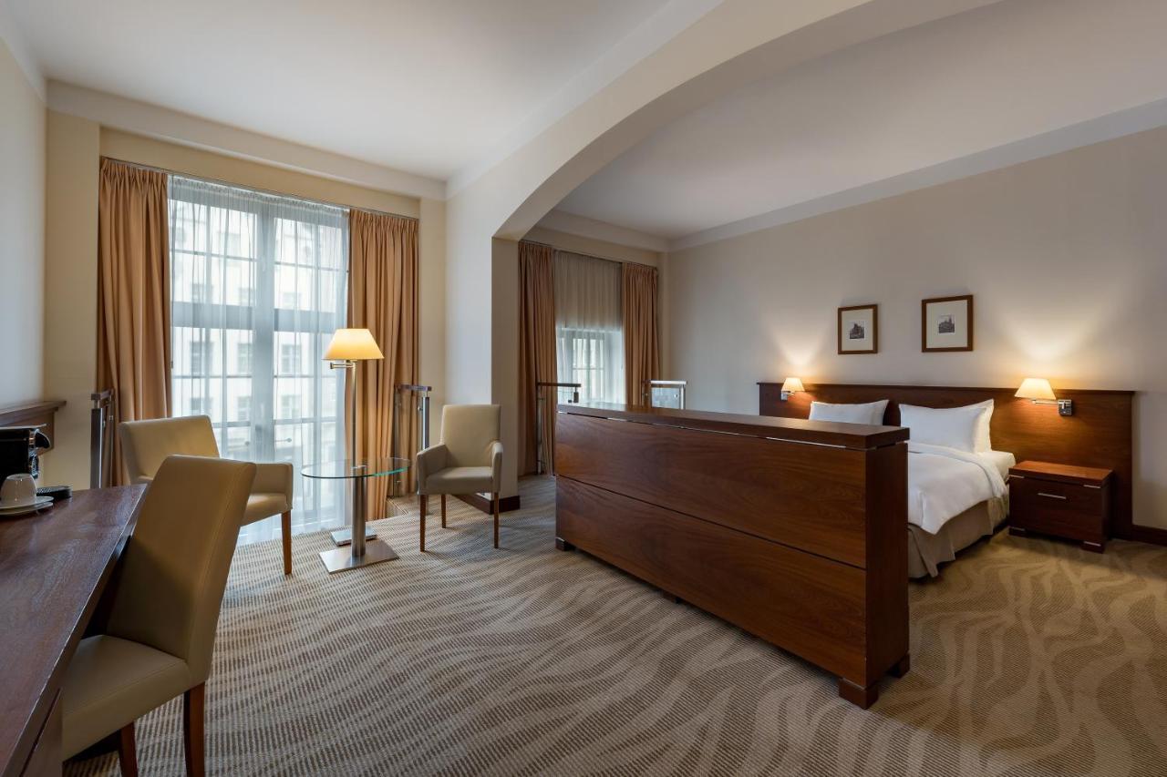 فندق راديسون بلو، غدانسك المظهر الخارجي الصورة The photo shows a well-furnished hotel room featuring a large bed with a wooden headboard positioned in the center. There are two chairs and a small table on one side, creating a cozy seating area. Near the window, which is covered with light curtain