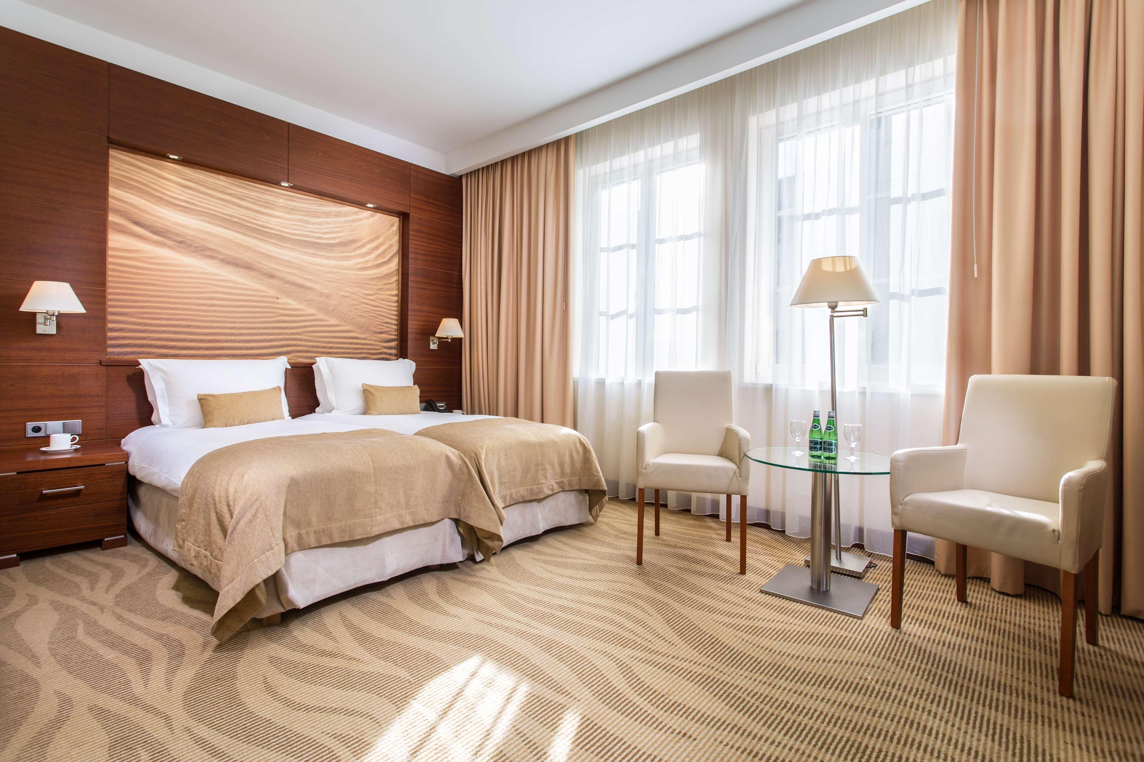 فندق راديسون بلو، غدانسك المظهر الخارجي الصورة The photo shows a well-decorated hotel room featuring two twin beds with beige linens and pillows. The walls have a warm, wooden paneling accent, and large windows are dressed with light curtains allowing natural light to fill the space. There are tw