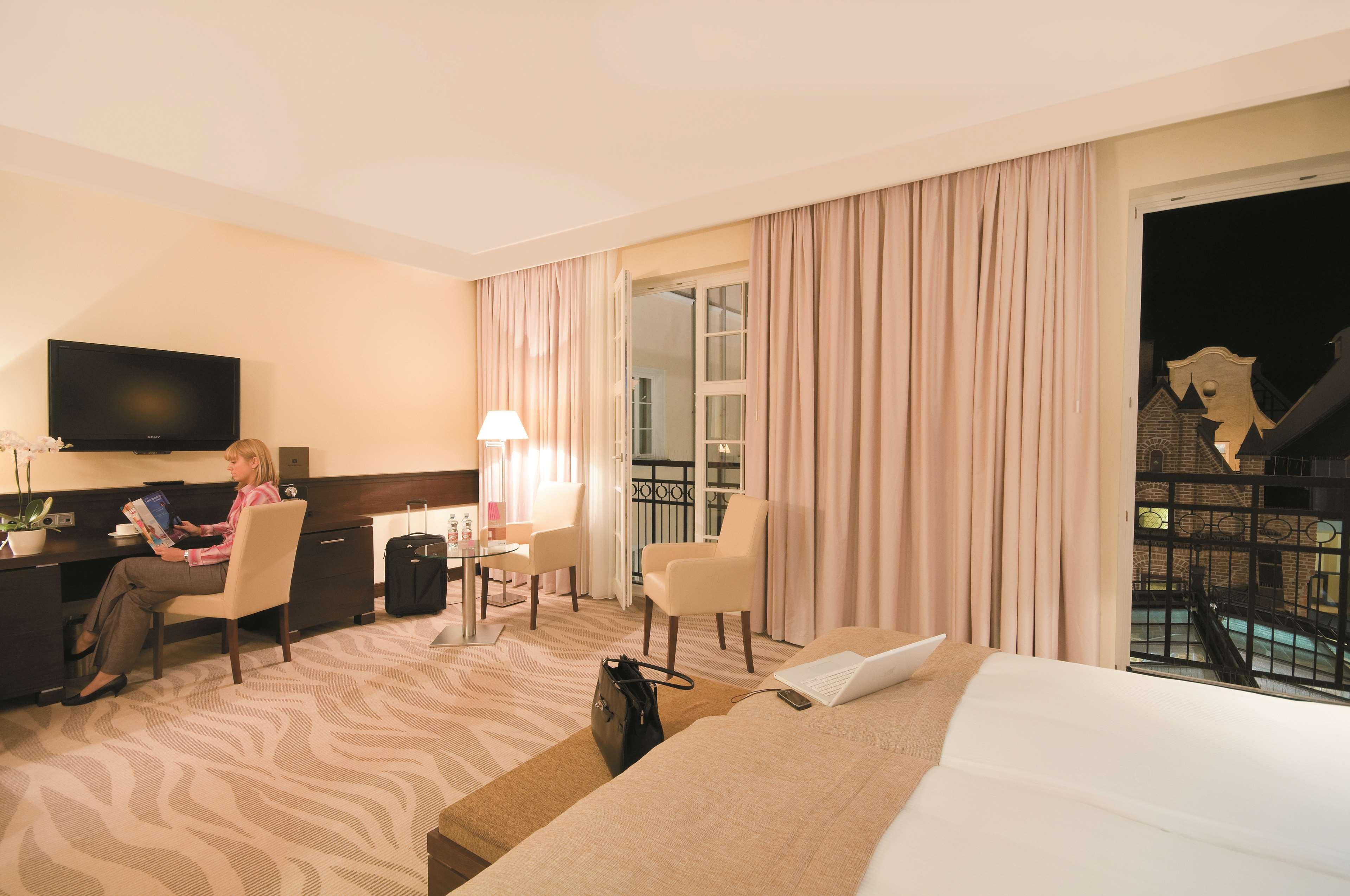 فندق راديسون بلو، غدانسك المظهر الخارجي الصورة The photo shows an interior view of a hotel room. On one side, there is a woman sitting at a desk with a laptop, working or using a computer. The room features a large bed with light-colored bedding and a patterned carpet. There are two chairs positi