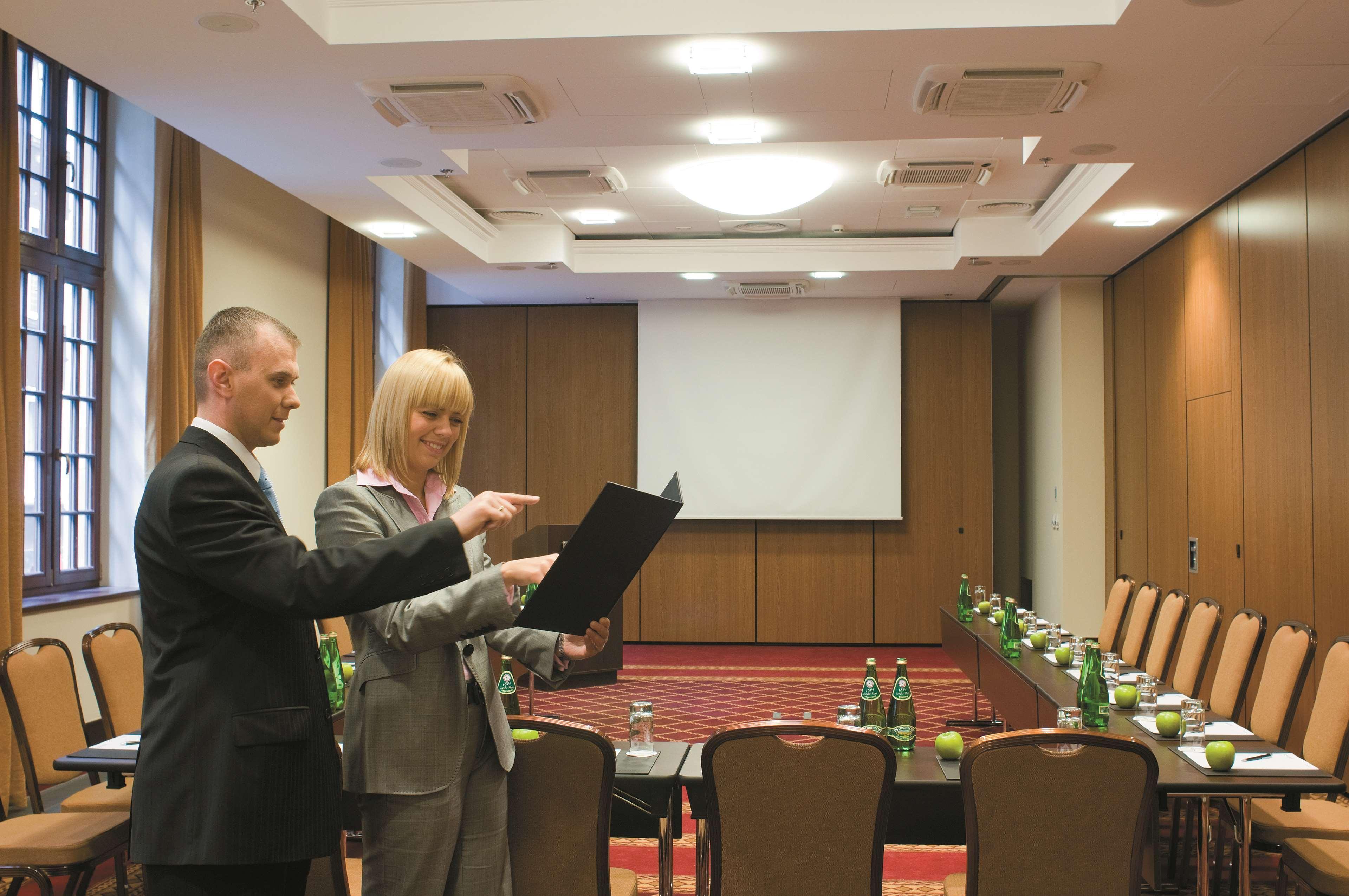 فندق راديسون بلو، غدانسك المرافق الصورة The image shows a professional meeting room set up for a conference or presentation. There are two people in formal attire engaged in discussion; one is holding a document or folder while the other is pointing at it. The room features a large screen 