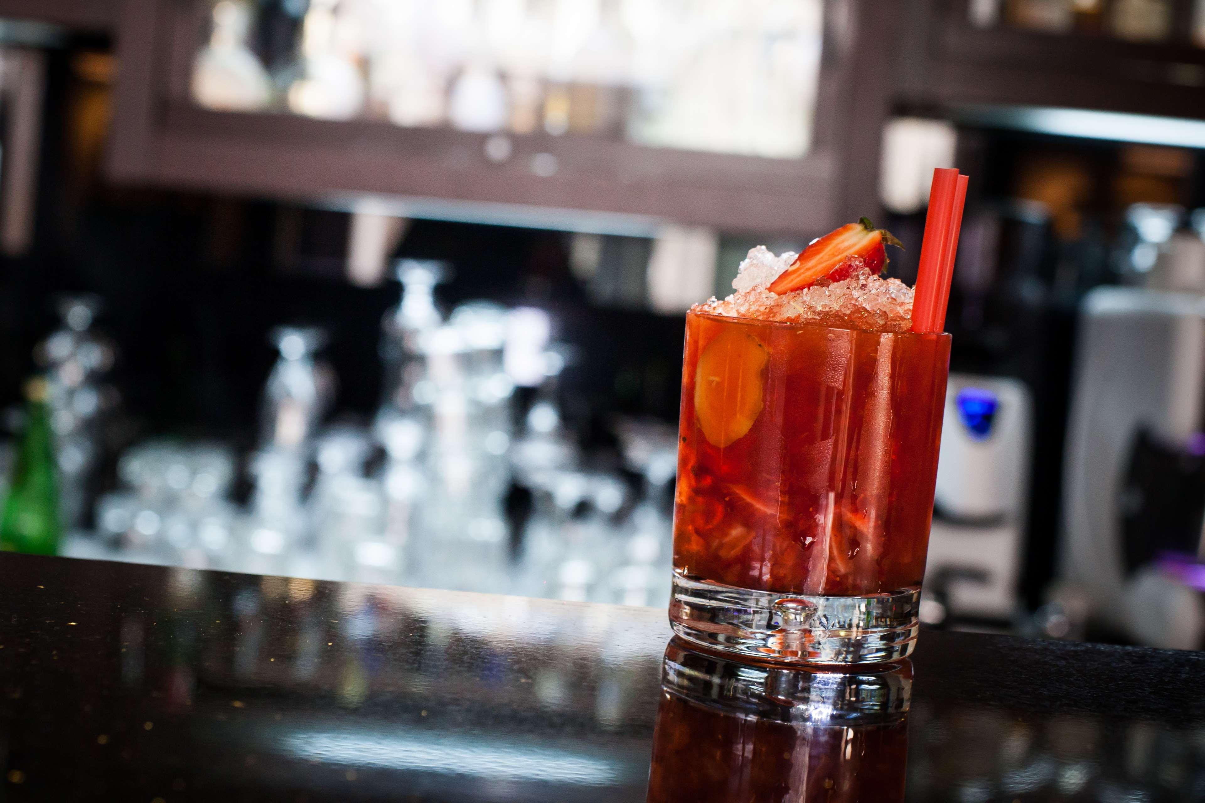 فندق راديسون بلو، غدانسك المظهر الخارجي الصورة The photo shows a glass of a colorful cocktail placed on a dark surface, likely a bar countertop. The drink is garnished with slices of fruit, possibly strawberries, and is filled with ice. In the background, there are blurred outlines of other glass