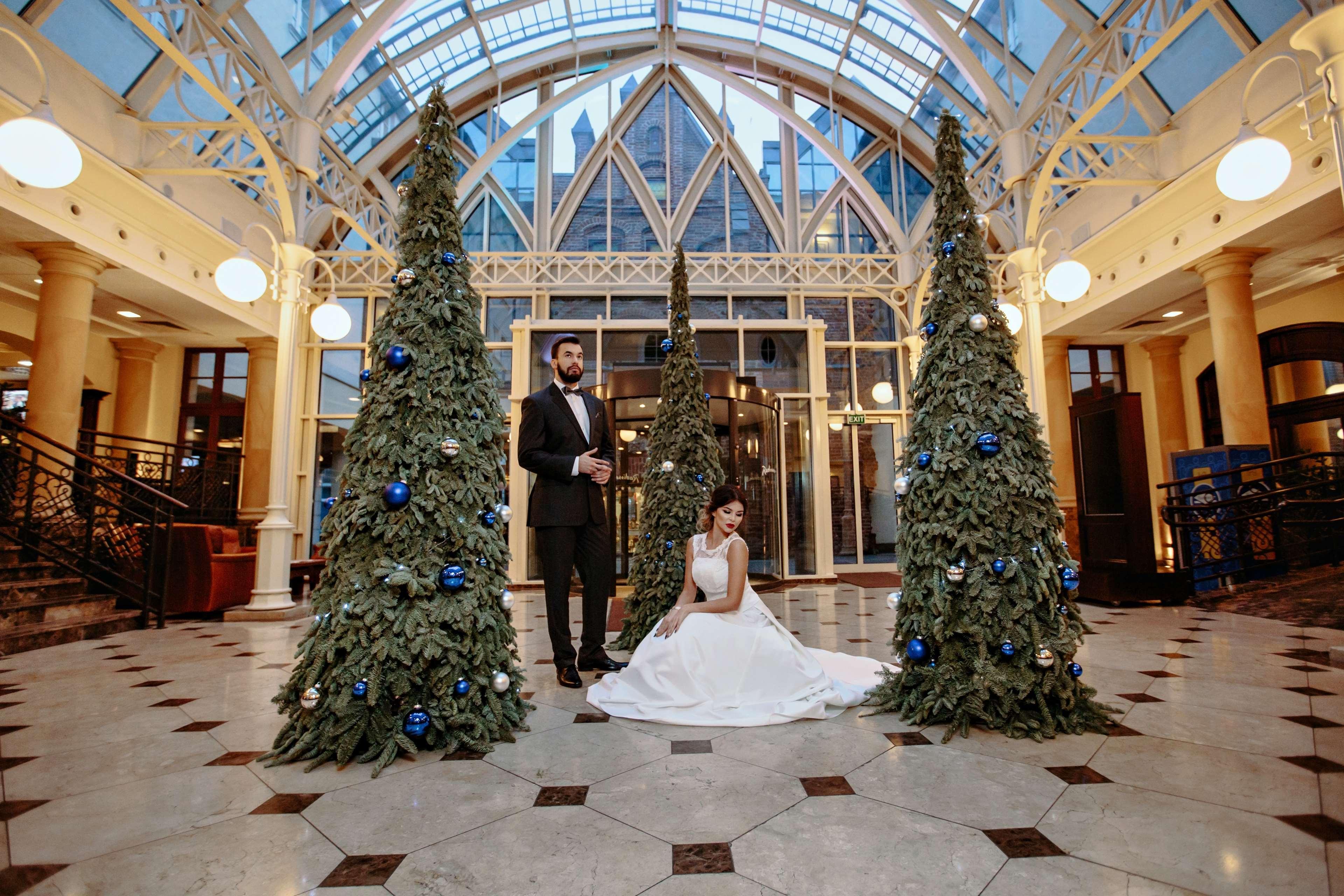 فندق راديسون بلو، غدانسك المظهر الخارجي الصورة The photo shows a groom in a tuxedo standing next to a bride in a white wedding dress. They are positioned in a beautifully decorated indoor space, which features large glass windows and elegant architectural details. Surrounding them are three green