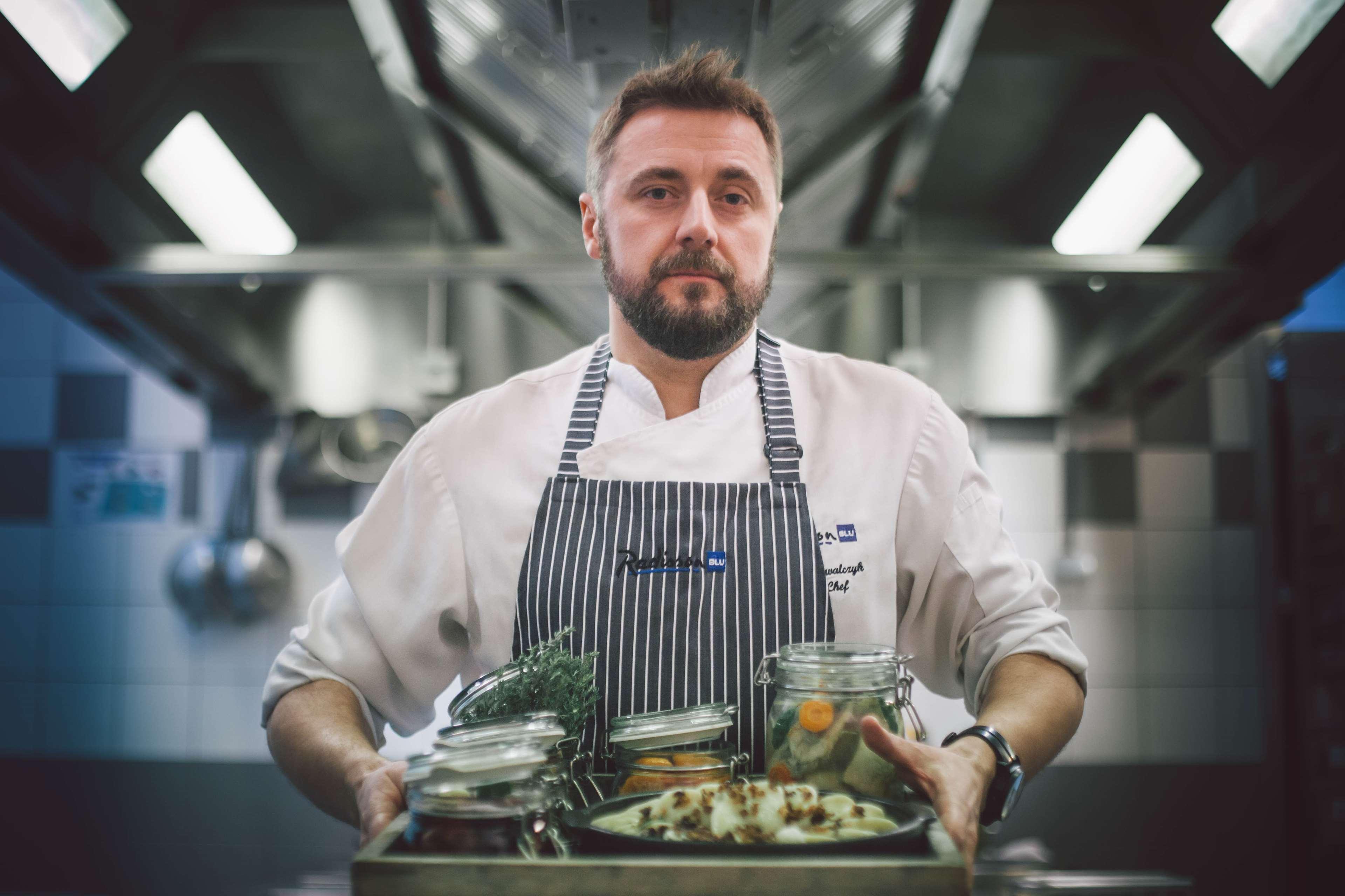 فندق راديسون بلو، غدانسك المظهر الخارجي الصورة The photo depicts a chef in a professional kitchen setting. He is wearing a white chef's coat with a striped apron and has a focused expression. He is holding a wooden tray filled with various jars and containers that likely contain ingredients for c