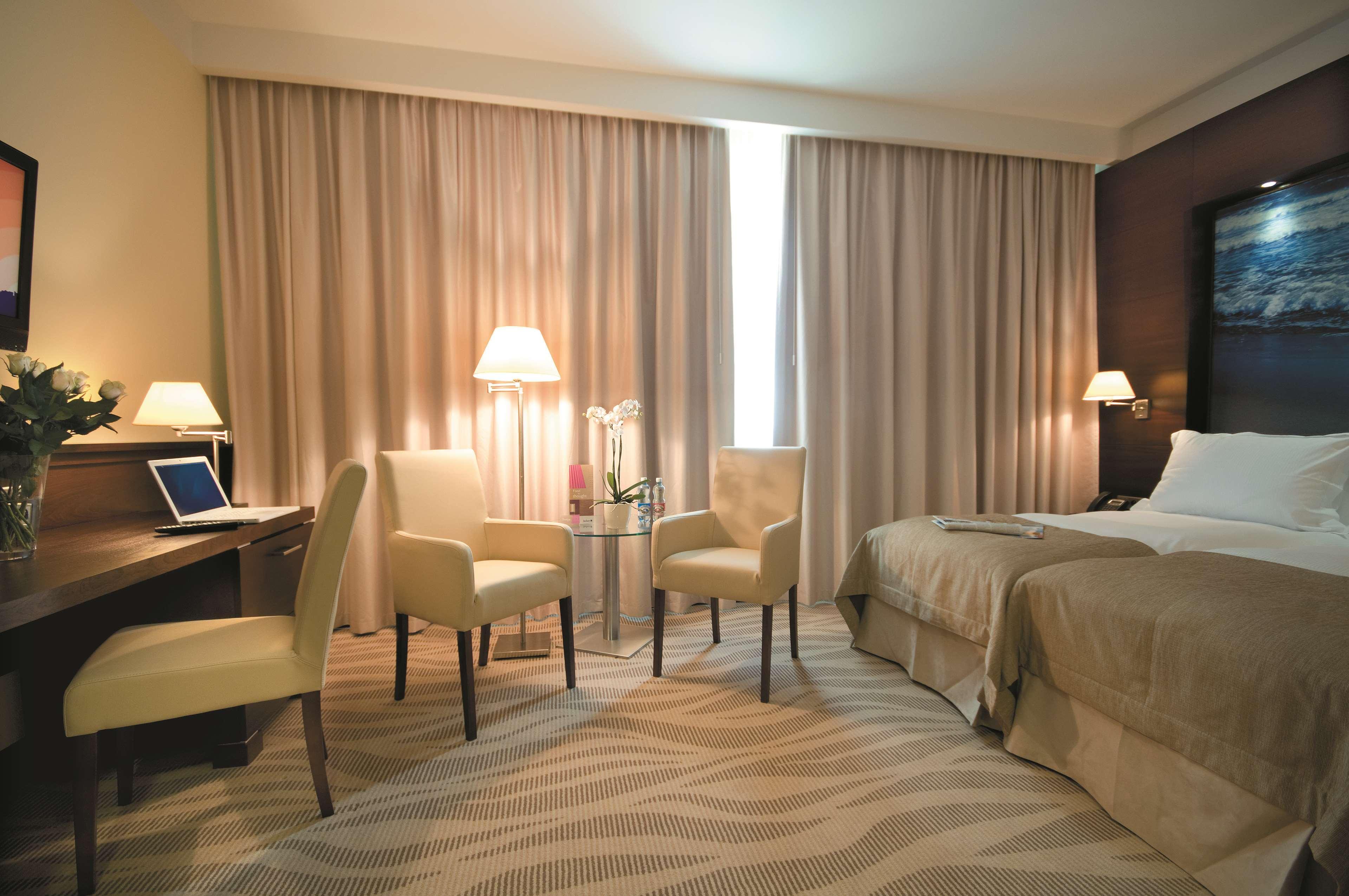فندق راديسون بلو، غدانسك المظهر الخارجي الصورة The photo shows a hotel room that features a queen-sized bed with neutral-colored bedding. There are two chairs and a small glass table in front of the bed. A desk is situated to one side, likely for work or personal use. The room has two lamps provi