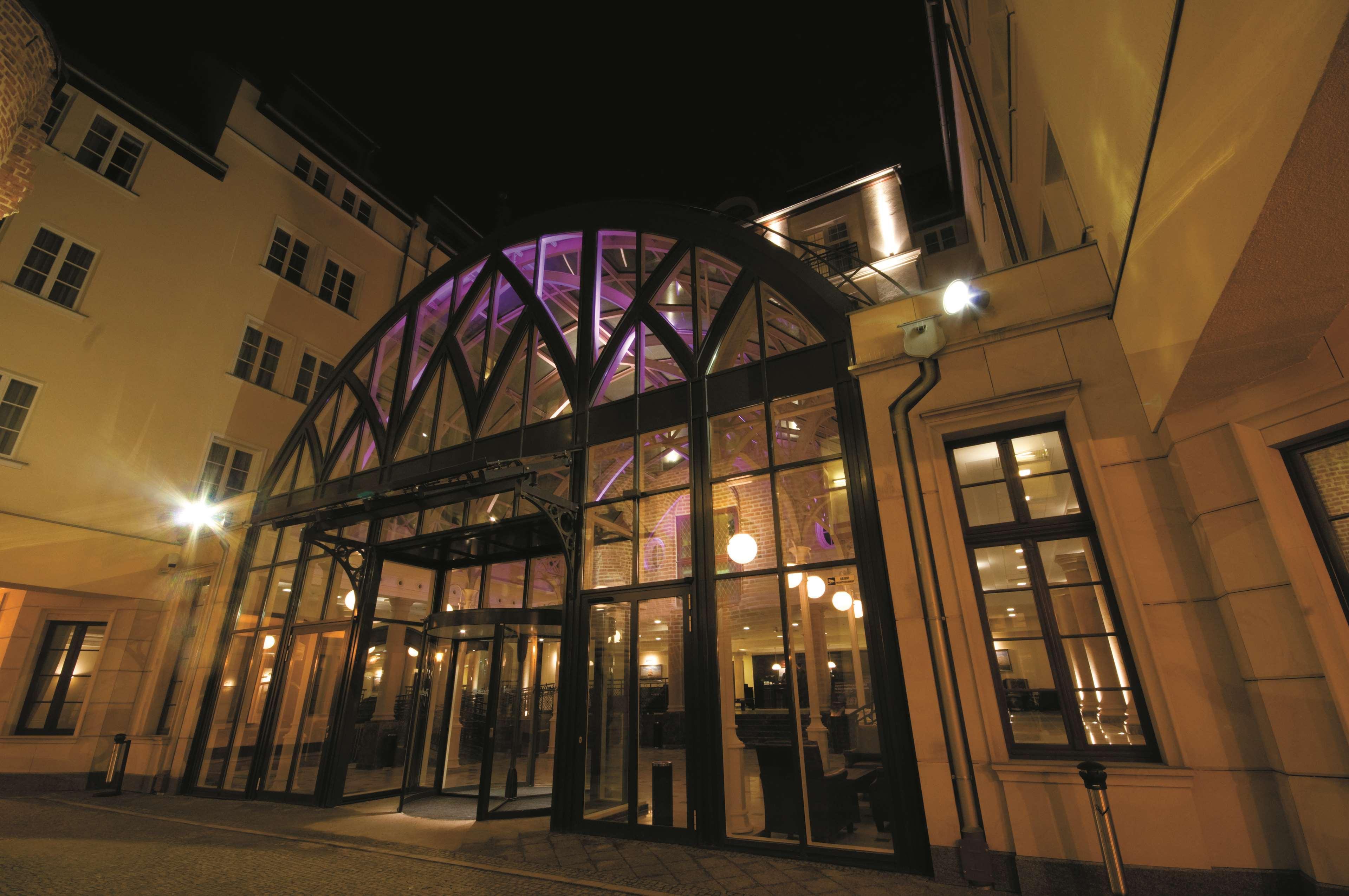 فندق راديسون بلو، غدانسك المظهر الخارجي الصورة The photo shows the entrance of a building at night. The structure features a large glass facade with an arched roof supported by a metal framework. The lights illuminate the entrance and the interior, creating a welcoming atmosphere. Soft purple lig