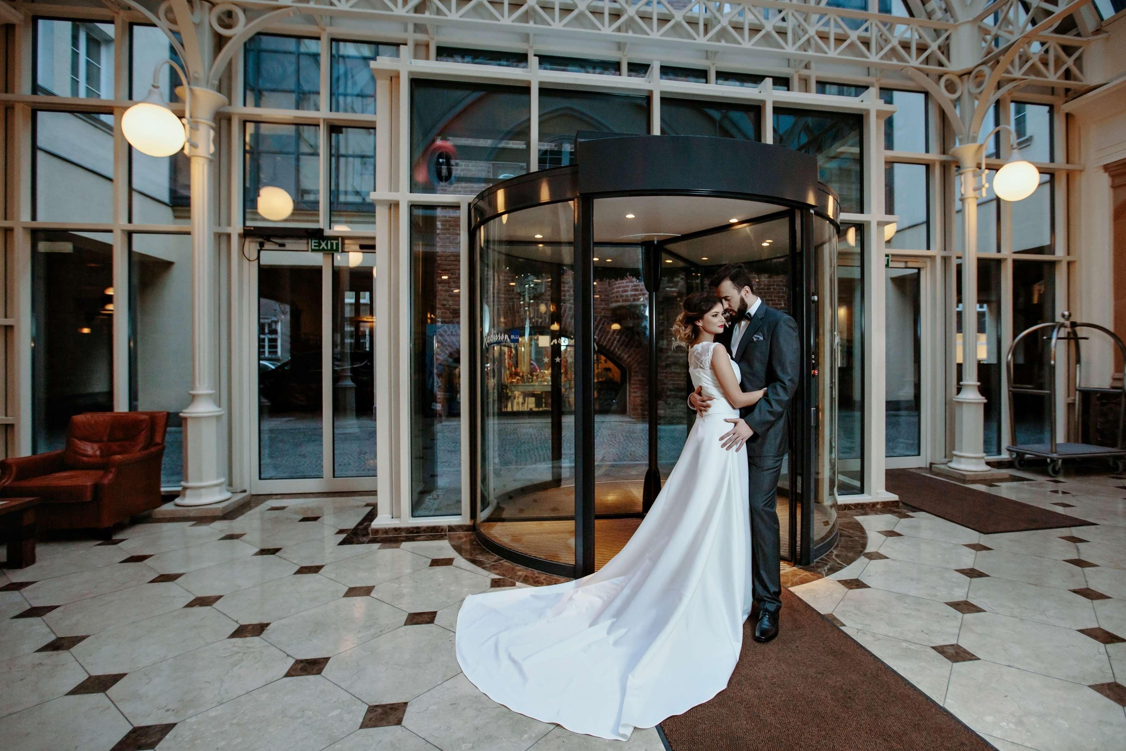 فندق راديسون بلو، غدانسك المظهر الخارجي الصورة The photo shows a couple in formal attire standing outside a building, likely a hotel or venue. They appear to be newlyweds, with the woman in a beautiful white wedding gown and the man in a suit. They embrace each other tenderly in front of a glass 