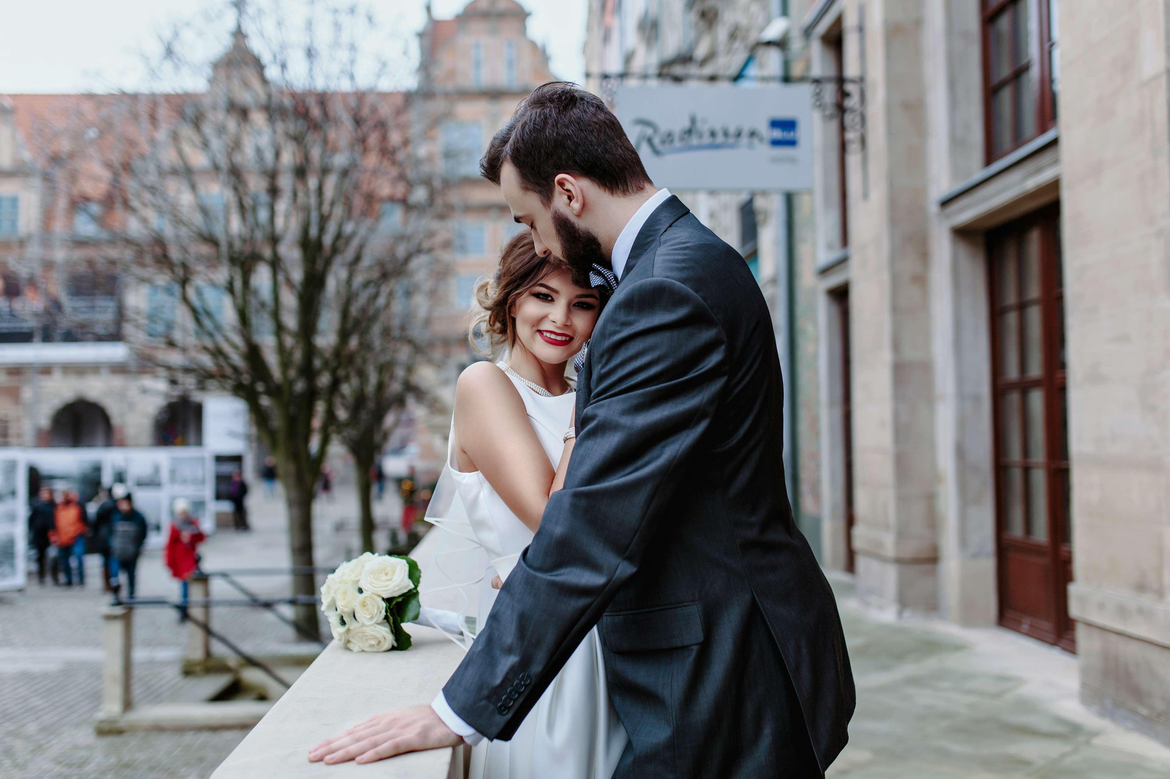 فندق راديسون بلو، غدانسك المظهر الخارجي الصورة The photo shows a couple embracing in an outdoor setting. The woman, dressed in a white wedding gown, is smiling and appears to be enjoying the moment. The man, wearing a dark suit, leans in toward her, suggesting a romantic atmosphere. In the backgr