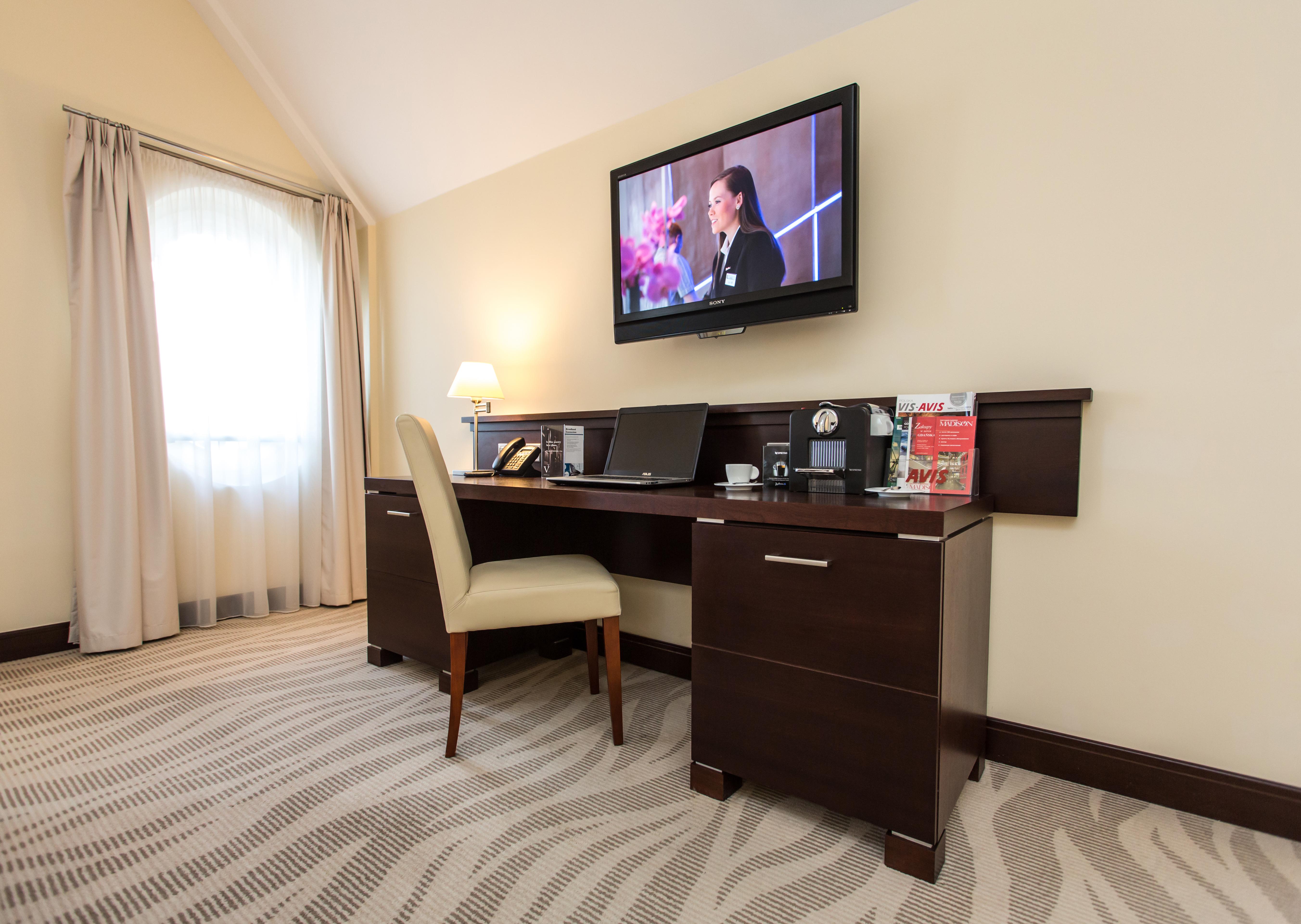 فندق راديسون بلو، غدانسك المظهر الخارجي الصورة The photo depicts a corner of a room featuring a stylish desk with a chair and a television mounted on the wall above. The desk has various items on it, possibly including a telephone, clock, and some decorative elements or stationery. There is a lam