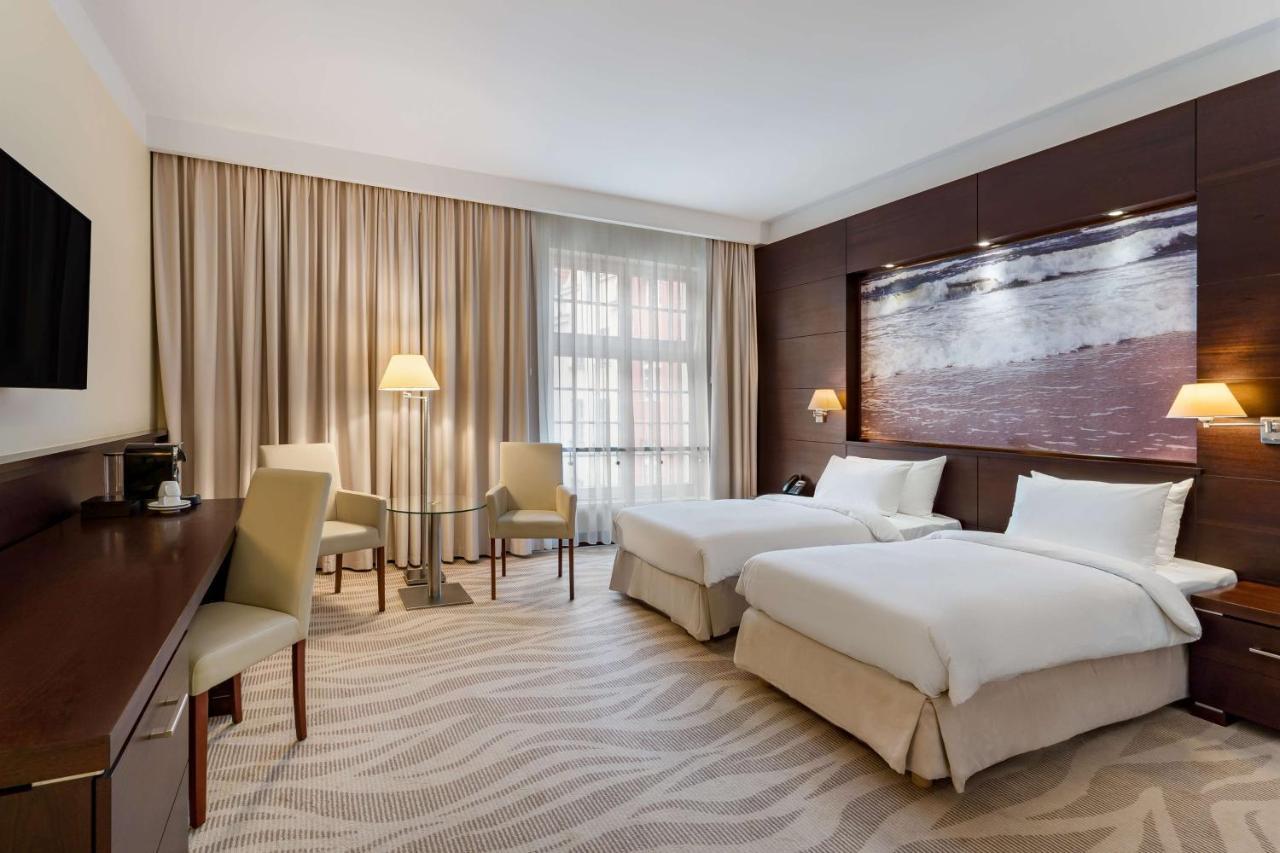 فندق راديسون بلو، غدانسك المظهر الخارجي الصورة The photo shows a neatly arranged hotel room featuring two beds with white bedding. There is a large window covered with light curtains, allowing natural light to enter the space. In one corner, there are two chairs and a small table, creating a cozy