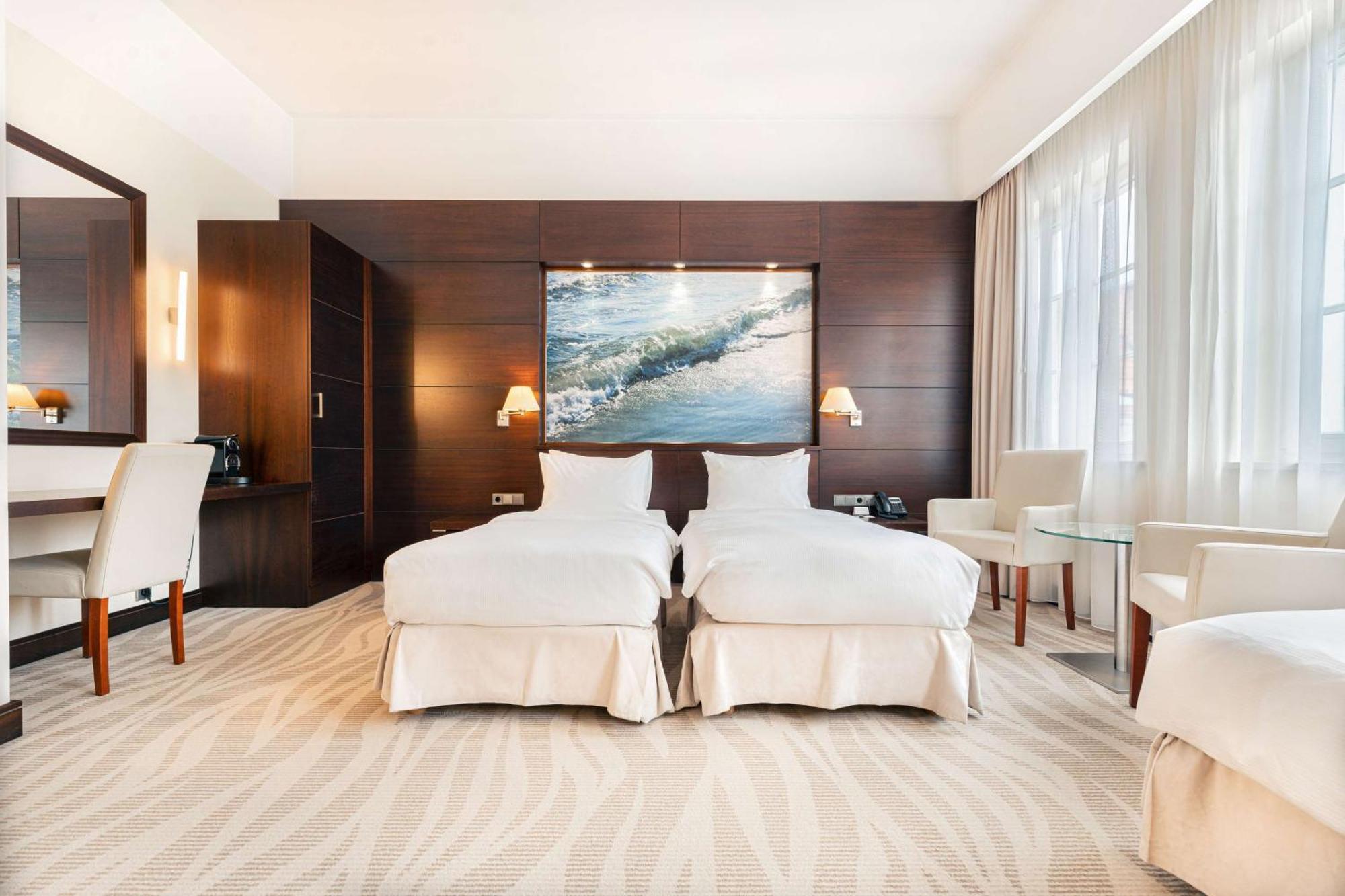 فندق راديسون بلو، غدانسك المظهر الخارجي الصورة The photo shows a modern hotel room featuring two twin beds dressed in white bedding. The room has a warm, elegant design with wooden cabinetry on one wall and a large piece of artwork depicting a landscape or seascape above the beds. There are two b