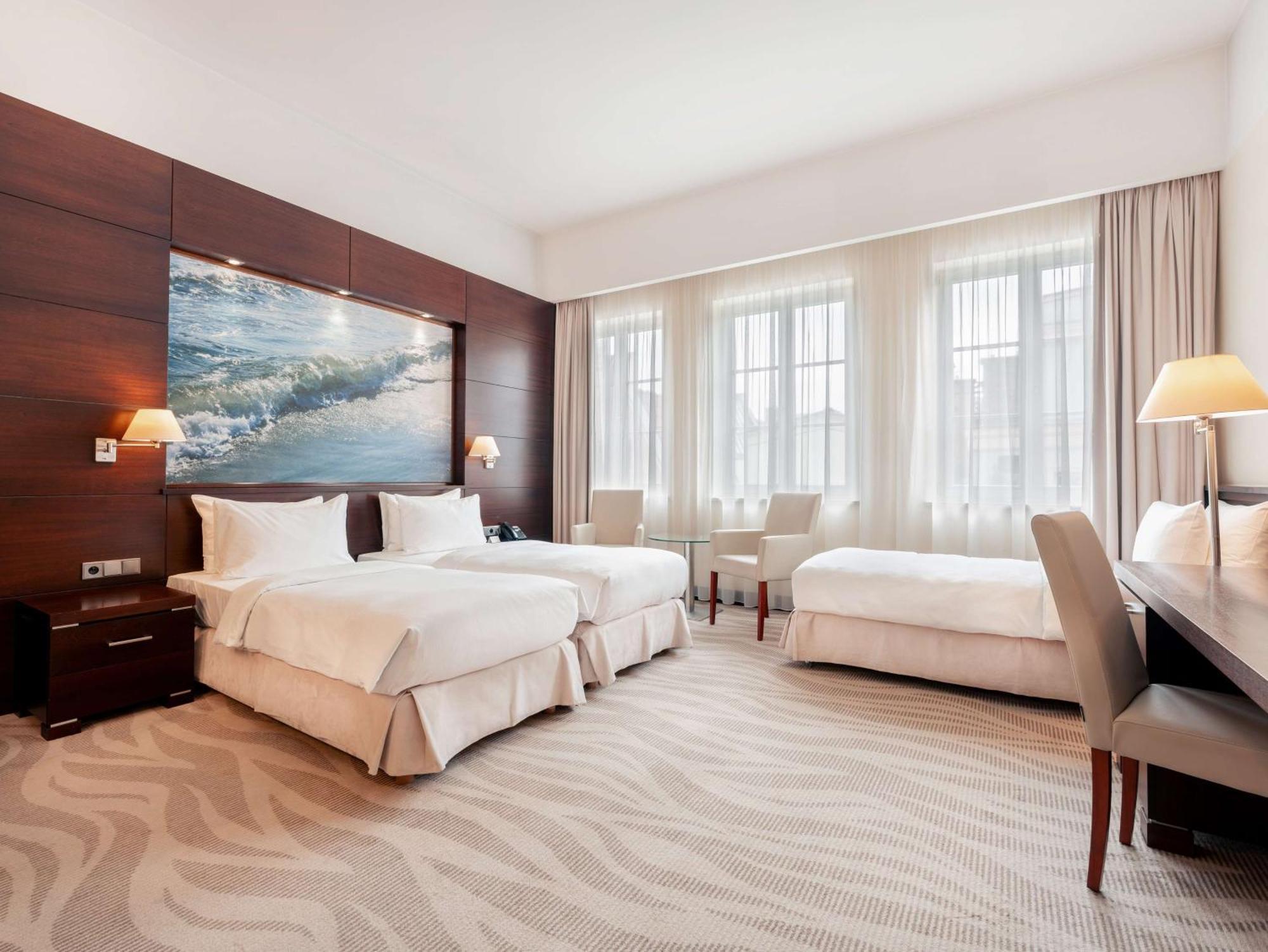 فندق راديسون بلو، غدانسك المظهر الخارجي الصورة The photo depicts a modern hotel room featuring two twin beds with white bedding, arranged against a wooden wall that includes a large artwork of ocean waves. The room is brightened by natural light streaming through three large windows, which are ad