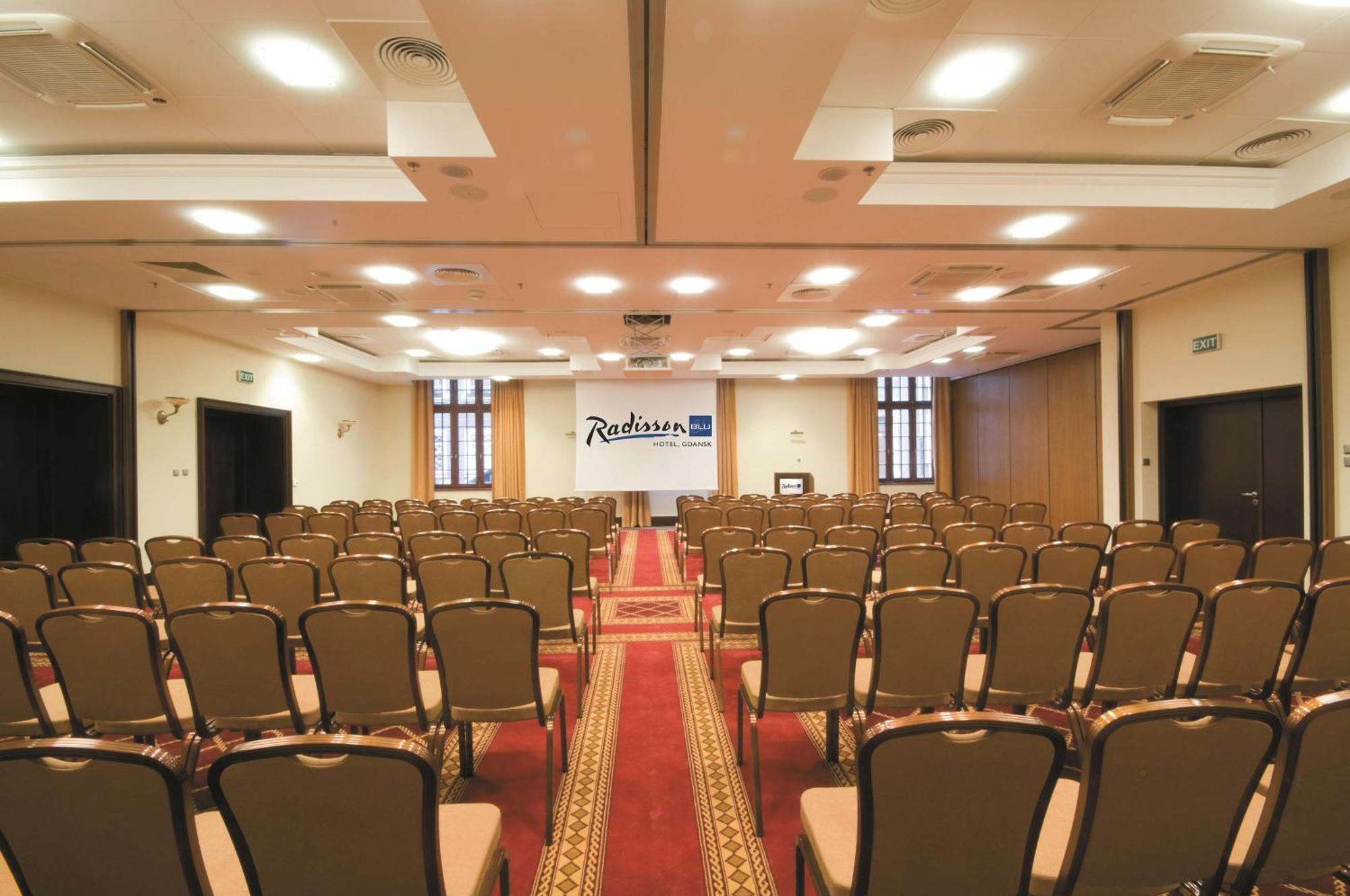 فندق راديسون بلو، غدانسك المظهر الخارجي الصورة The photo shows the interior of a conference room or event space. It features rows of brown chairs arranged neatly, facing towards the front where a stage or presentation area is set up. There is a carpet running down the center of the room, and the 
