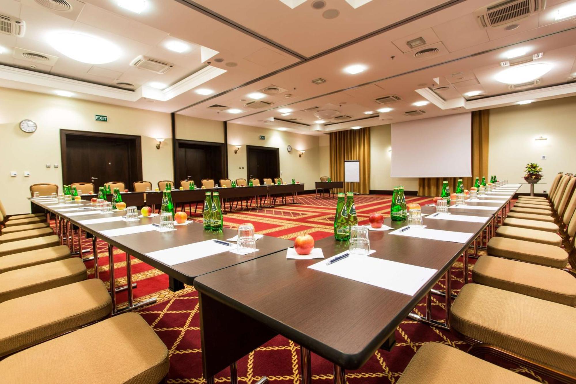 فندق راديسون بلو، غدانسك المظهر الخارجي الصورة The photo shows a conference room set up for a meeting or seminar. Long tables are arranged in a U-shape, surrounded by beige chairs. Each table has green glass bottles of water, small glasses, and apples placed on it. There are also sheets of paper 