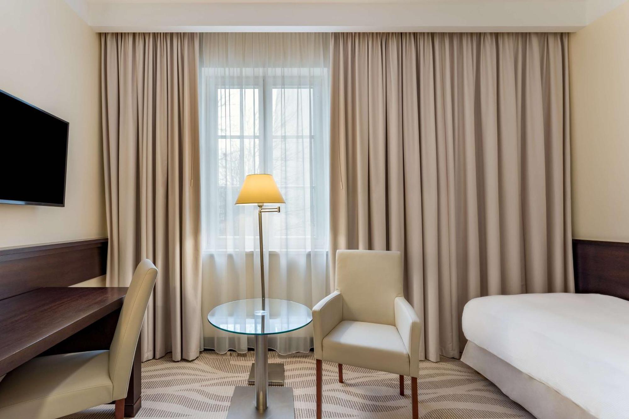 فندق راديسون بلو، غدانسك المظهر الخارجي الصورة The photo shows a hotel room that features a light and neutral color scheme. There is a window covered with sheer curtains, allowing natural light to enter the space. To the right, there is a bed with a white bedspread. 

In the center, there is a sm