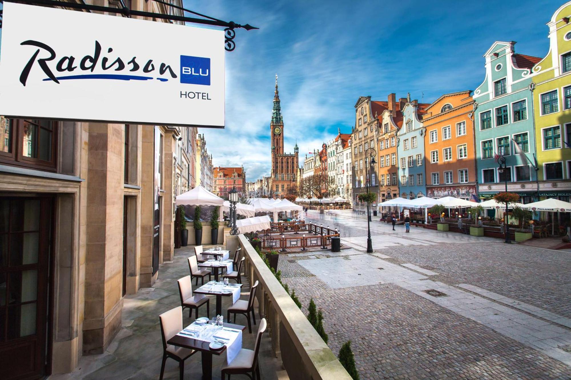 فندق راديسون بلو، غدانسك المظهر الخارجي الصورة The photo shows a view of the Radisson Blu Hotel, which is situated in a vibrant, picturesque area. The image features an outdoor seating arrangement with tables and chairs, likely part of a restaurant or café. In the background, colorful historic bu