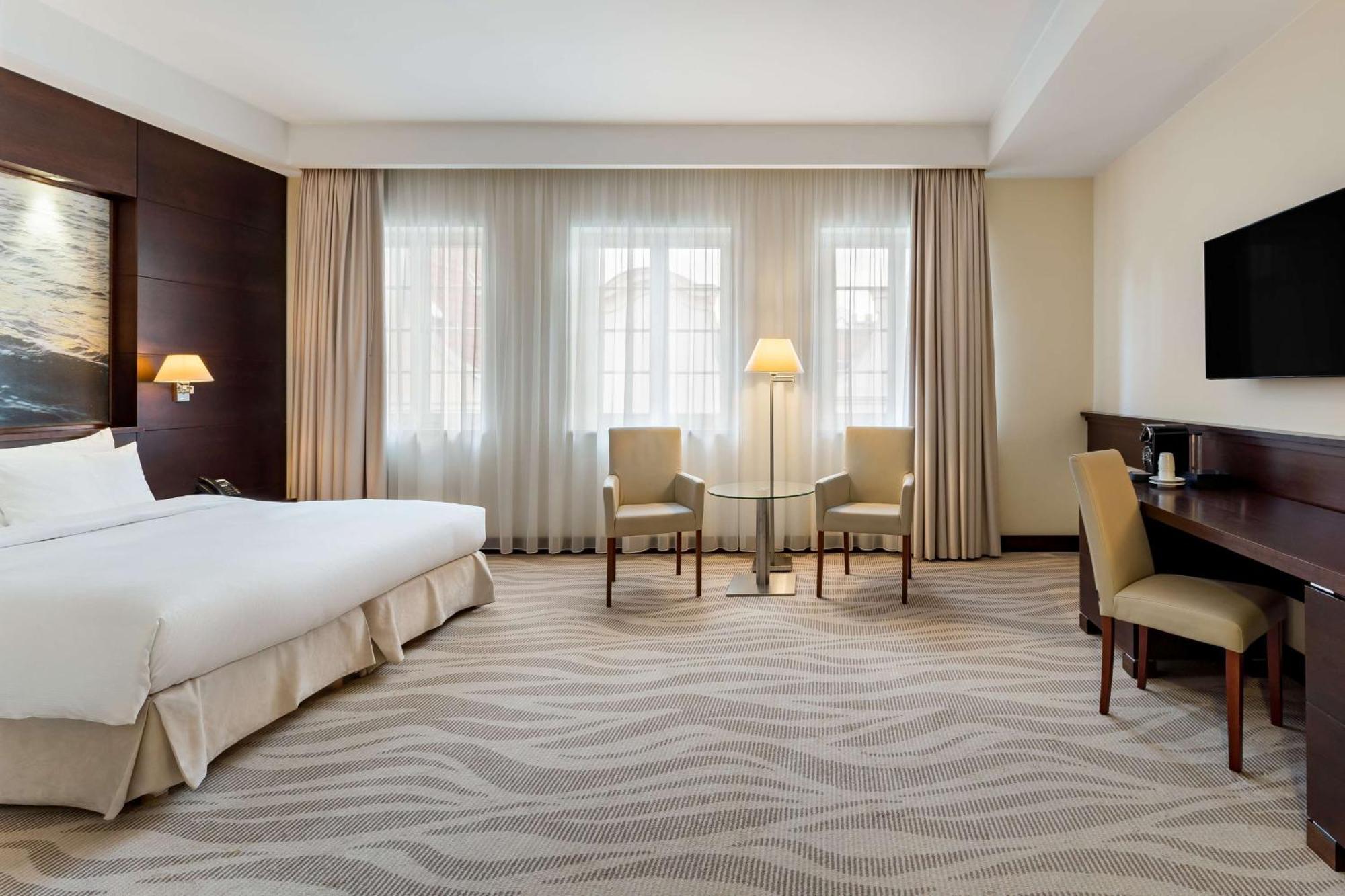 فندق راديسون بلو، غدانسك المظهر الخارجي الصورة The photo shows a hotel room featuring a modern and clean design. There's a large bed with white bedding positioned against a wall, accompanied by bedside tables. Two armchairs and a small side table are set near a lamp in front of large windows that