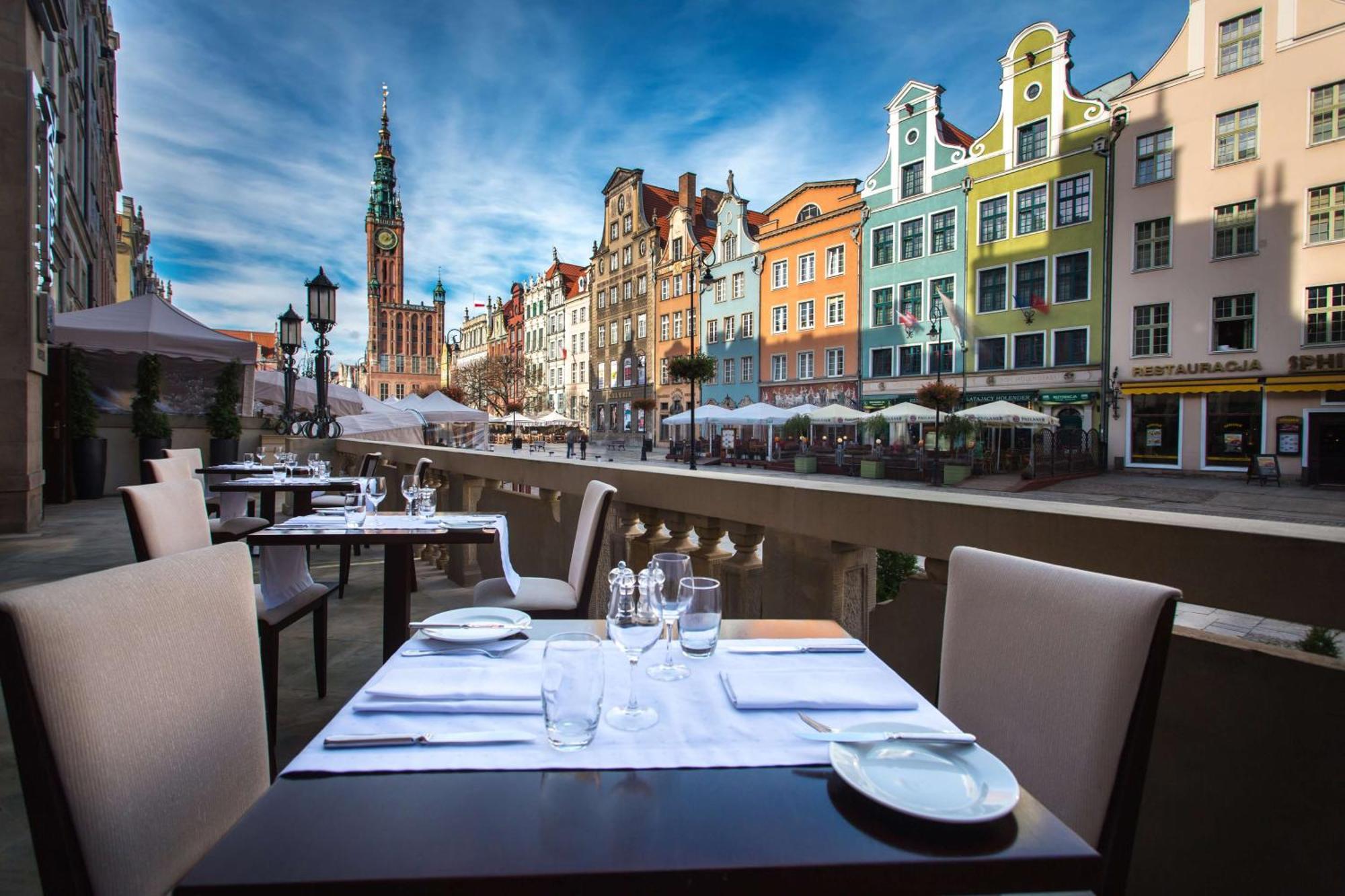 فندق راديسون بلو، غدانسك المظهر الخارجي الصورة The photo shows an outdoor dining area with elegantly set tables overlooking a picturesque street lined with colorful buildings. In the background, there is a tall clock tower, suggesting a historic or charming town. The sky is bright with some cloud