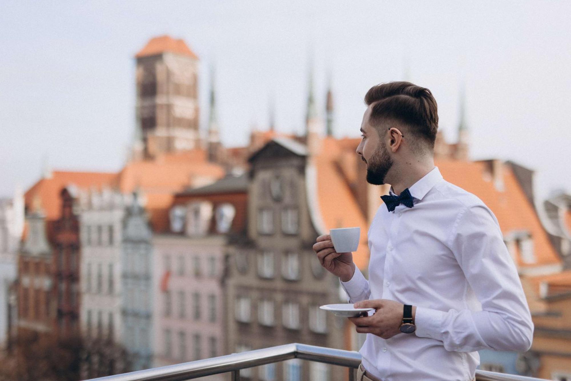 فندق راديسون بلو، غدانسك المظهر الخارجي الصورة The photo features a man standing on a balcony, holding a cup of coffee or tea in one hand while resting a saucer in the other. He is dressed in a white shirt with a bow tie. The background reveals a picturesque view of an old town, showcasing buildi